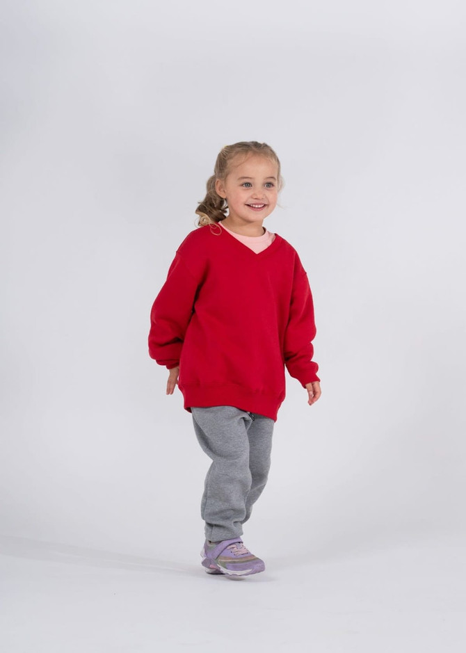 A child in a red v-neck fleece jumper and grey pants, smiling and walking against a plain background.