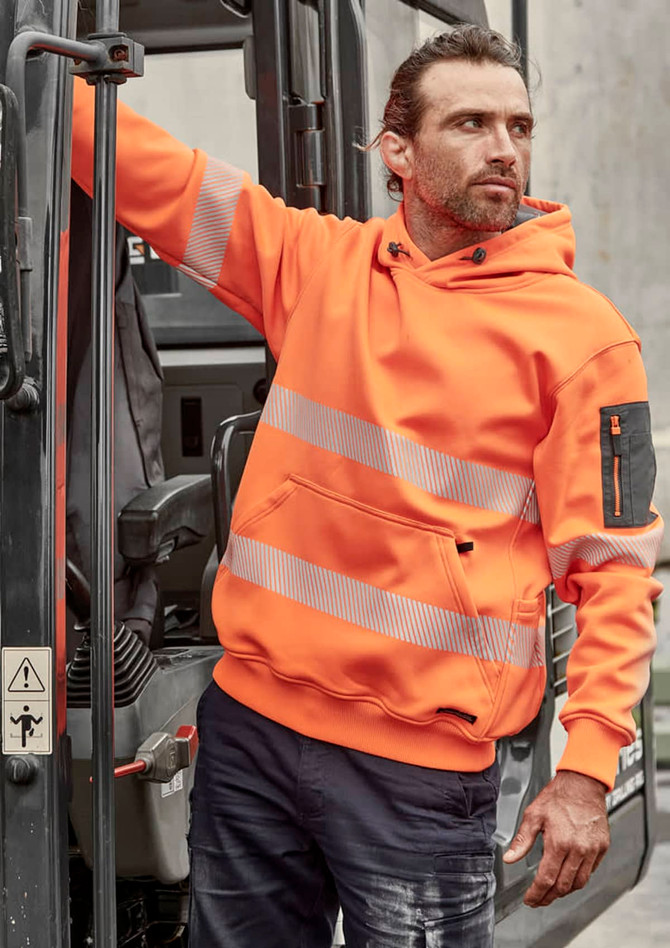 An orange water-resistant hoodie features segmented tape and a logo, worn by a model near machinery.