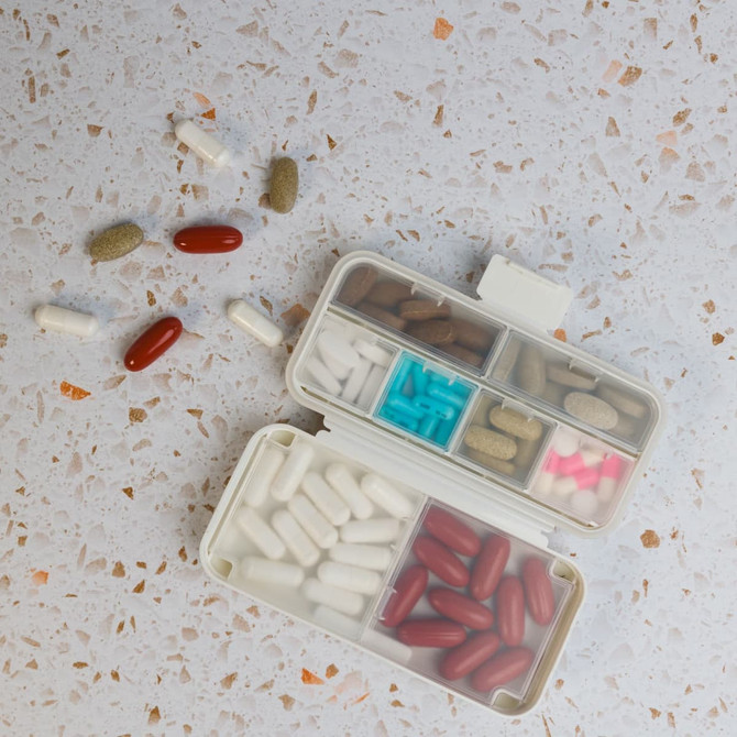 A compact pill organiser in white, with compartments for various coloured pills and capsules scattered around.