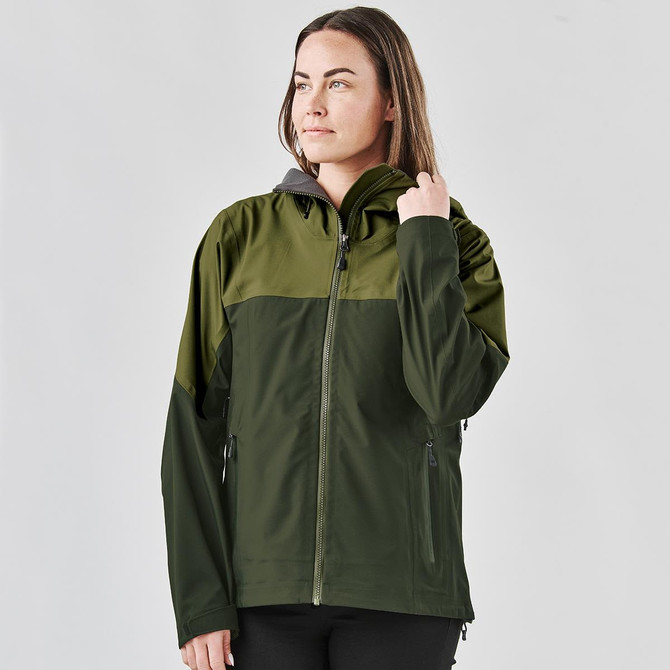 A woman models a green and dark green Women's Vertex Stormshell jacket with a hood. The jacket features a logo.