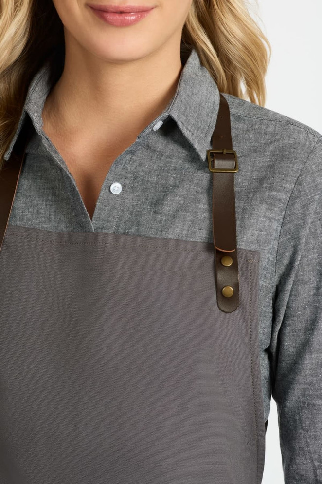 A ladies long sleeve button-up shirt in grey, featuring brown leather straps on an apron. It has a logo.