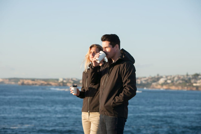 A couple wearing black soft shell jackets stands by the water, holding coffee cups and enjoying the scenery.