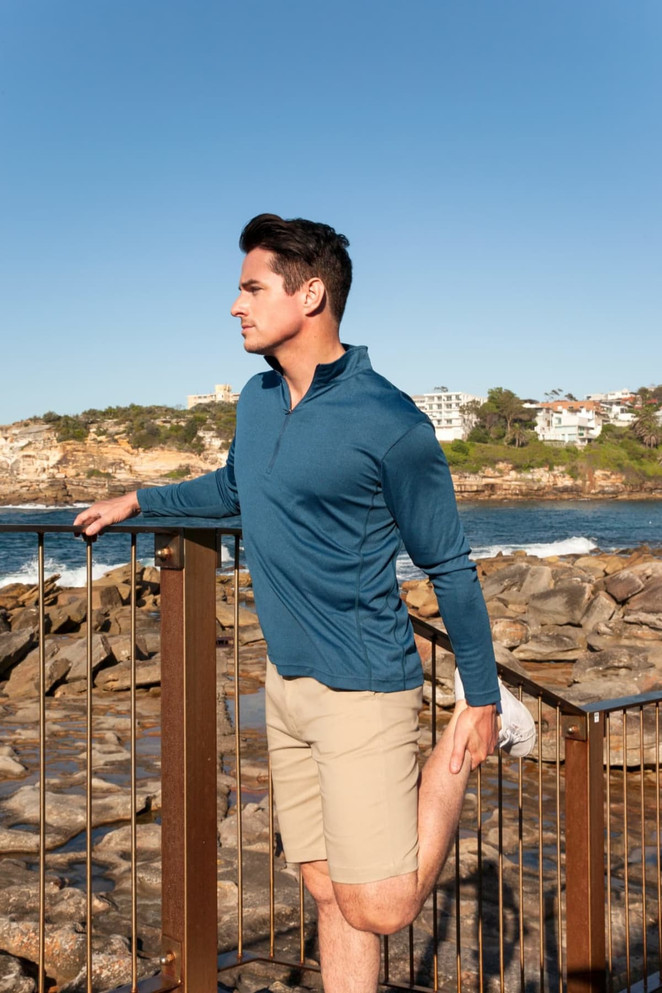 A man in a blue long-sleeve top and beige shorts stretches by the water, with rocky terrain in the background.