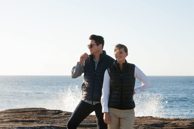 A black puffer vest worn by a man and a woman, standing by the water with a coastal background.