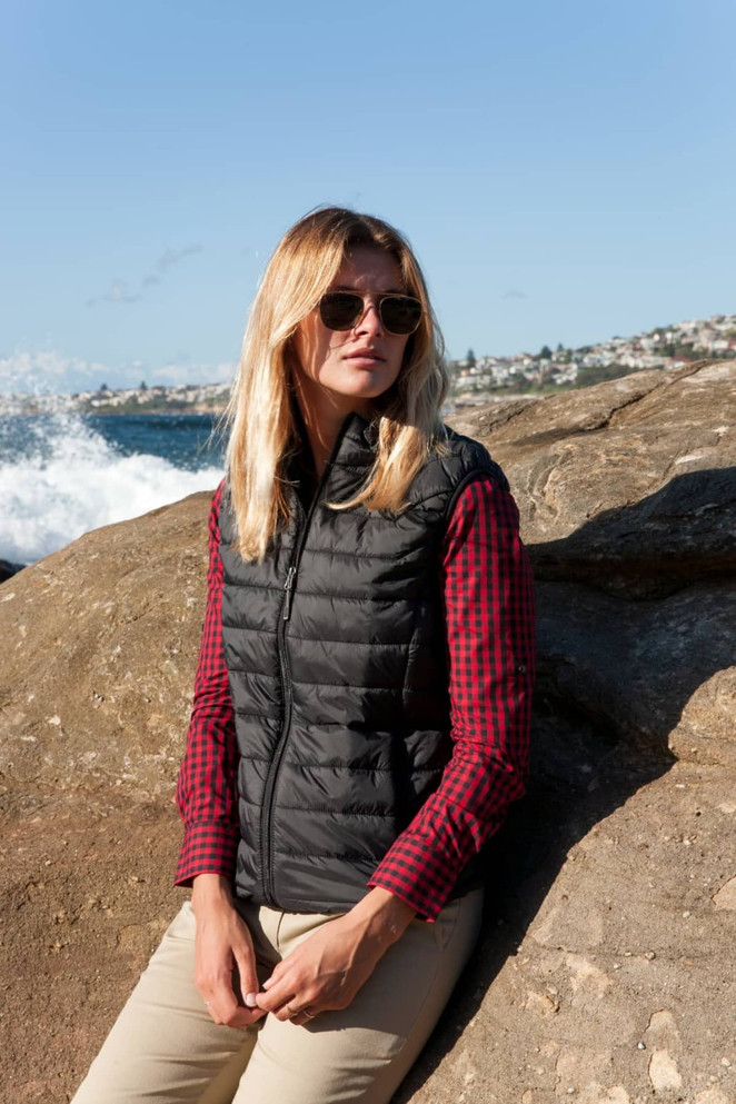 A black puffer vest worn by a woman with long blonde hair, sitting on a rock by the sea.