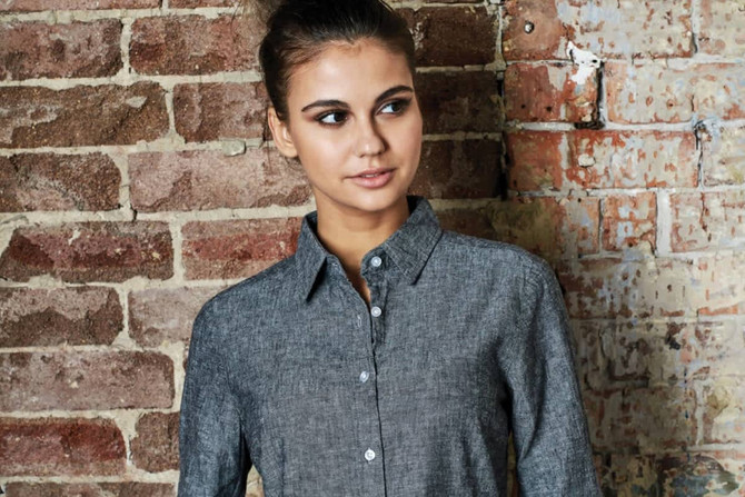A grey button-up long sleeve shirt is displayed against a brick wall, featuring a collared neckline and button closure.