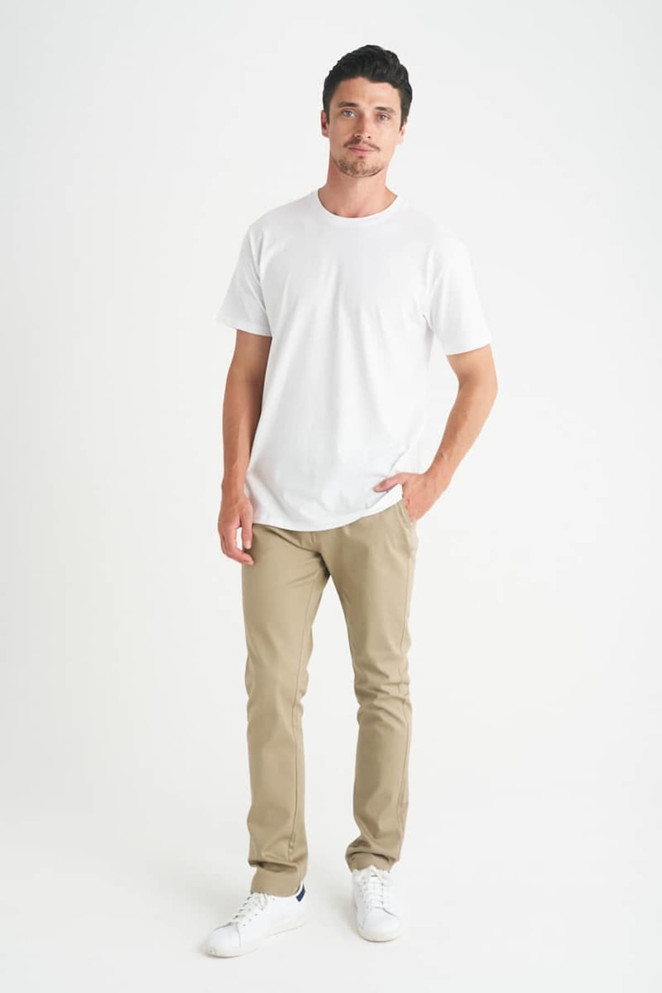 A man stands wearing a plain white t-shirt and beige pants, with one hand in his pocket.