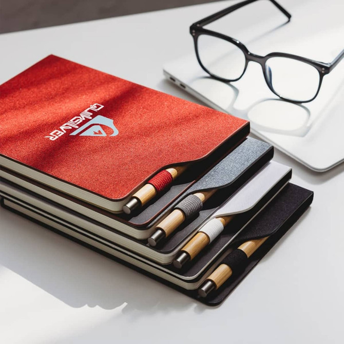 A set of A5 notebooks in red, black, white, and grey with matching pens, placed next to glasses and a laptop.