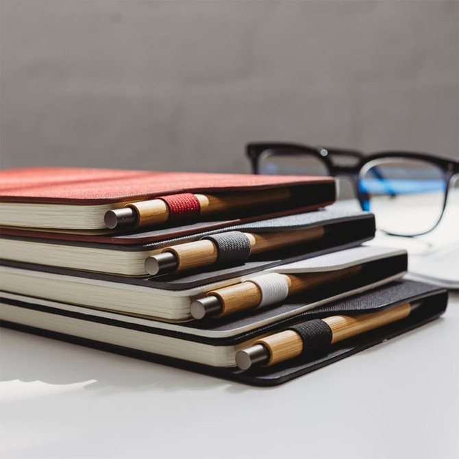 A set of RPET A5 notebooks in red, black, and white, featuring bamboo pens, stacked beside glasses on a desk.