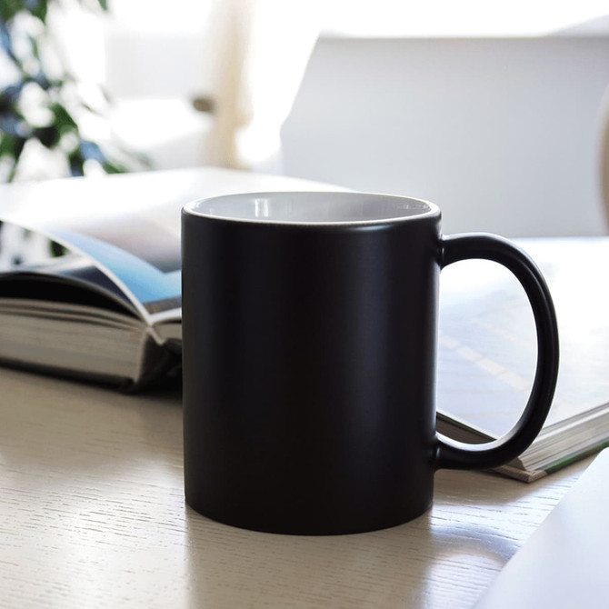 A ceramic mug in matte black with a white interior, placed on a table next to magazines. It features a handle and a logo.