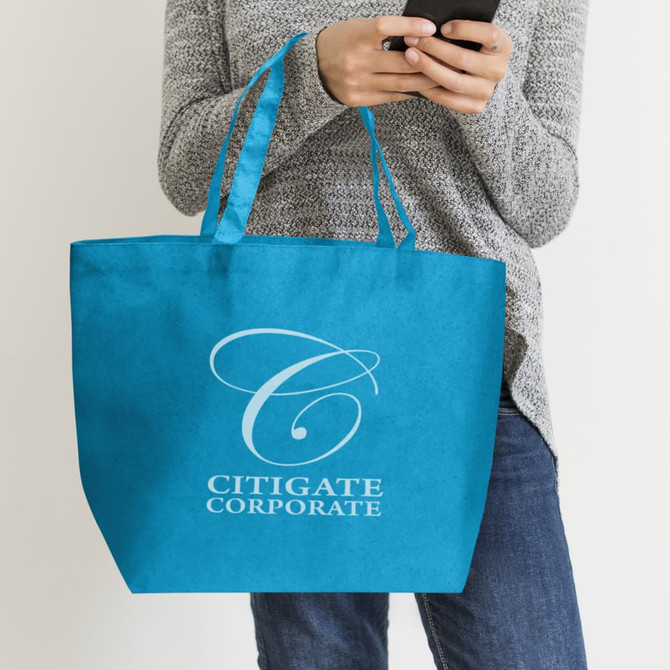 A blue tote bag with a logo, held by a person wearing a grey sweater and jeans, holding a mobile phone.