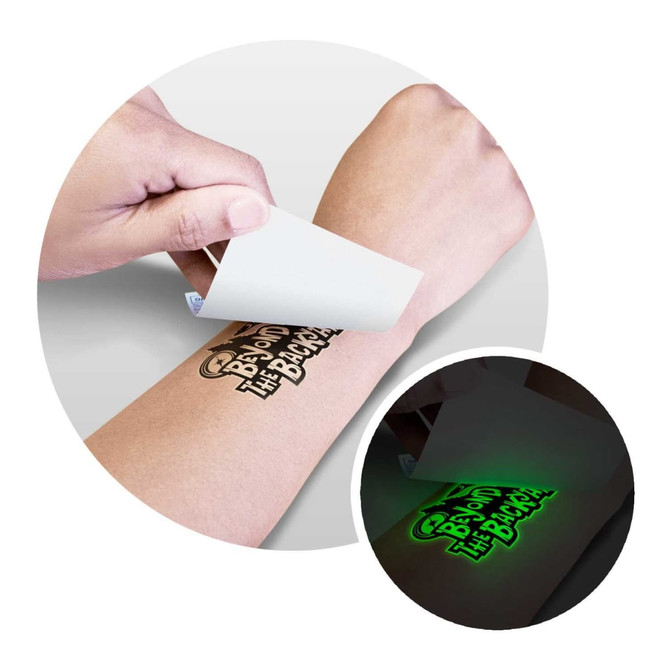 A glow-in-the-dark temporary tattoo being applied to a person's arm. The tattoo features bold black designs on a skin tone background.