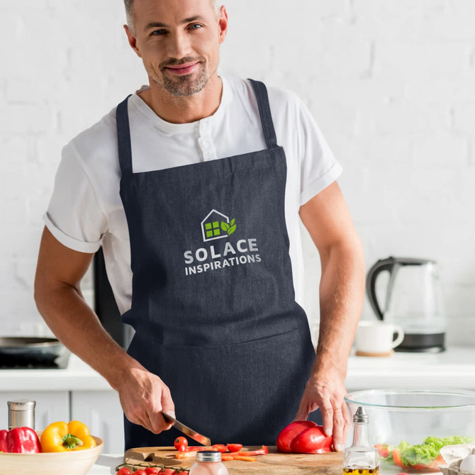 A man wearing a navy denim bib apron, chopping vegetables in a bright kitchen, featuring a logo on the apron.