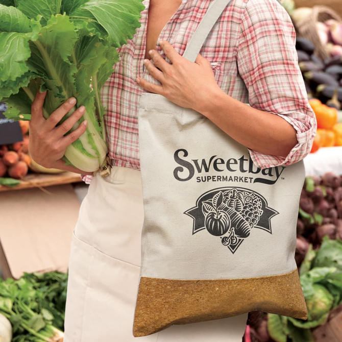 A neutral-coloured tote bag with a logo, held by a person carrying fresh produce in a market setting.