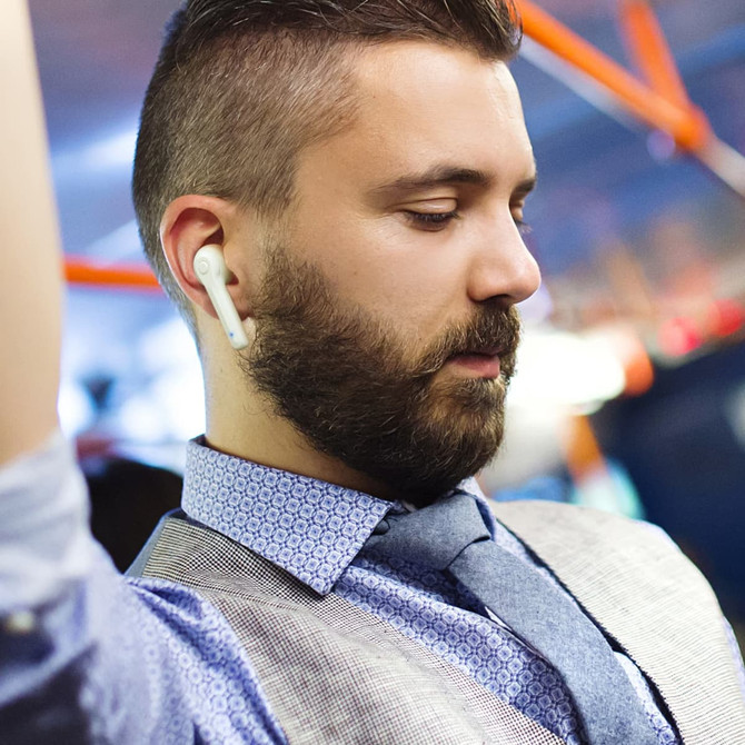 A man with a beard wearing a grey vest and tie, listening with white Bluetooth earbuds in a busy environment.