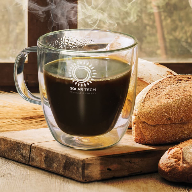 A clear double wall glass cup filled with dark coffee, placed next to fresh bread on a wooden surface.