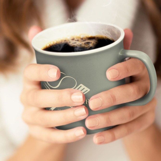 A green coffee mug held in hands, filled with steaming black coffee. The mug features a logo.