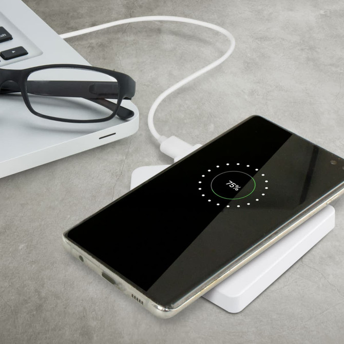 A white wireless charger with a smartphone displaying a charging symbol, next to glasses and a laptop.