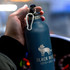 A drink bottle in matte blue with a silver top and a logo featuring a wolf, held by a hand.