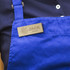 A blue apron features a magnetic wooden badge with a logo and name displayed prominently.