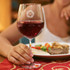 A wine glass with a decorative logo, filled with red wine, held by a person beside a plate of food.