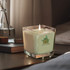 A green, square scented candle in a glass container with a logo, placed on a bedside table beside a book.