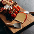 A wooden cheese board featuring cheese, cherry tomatoes, blueberries, and a set of metal cheese knives with wooden handles.