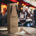A brown paper bag with a ribbon handle alongside a glass of red wine, set in a casual dining environment.