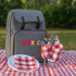 A grey picnic backpack rests on a red and white checkered picnic blanket, alongside utensils and napkins in glasses.