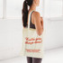 A calico tote bag in cream colour featuring a logo and text. A person holds it while wearing activewear.
