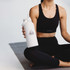 A drink bottle in white, featuring a logo, held by a person in activewear while seated on a yoga mat.