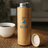 A drink bottle in bamboo finish with a silver lid and a logo. It sits on a woven surface near a cup.