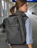 A grey backpack worn on the back of a person, set in a blurred airport environment. It features a logo.