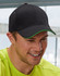 A black cap with a green peak, featuring a logo, worn by a smiling man in a bright yellow shirt.