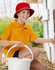 A red bucket hat sits on a boy wearing an orange polo shirt, holding a white bucket in a garden setting.