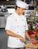 A chef wearing a white short sleeve jacket and a matching hat, chopping vegetables in a kitchen setting.