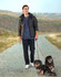 A man wearing a black sustainable parka jacket walks two dogs along a rural path, set against a hilly background.