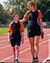 A black and orange Kids' TrueDry tri-colour S/S pique polo is worn by a child and an adult on a running track.