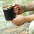A drink mug with a stainless steel rim and a black exterior, held by a person outdoors.