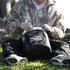 A black cooler bag is being opened by a person sitting on grass, wearing a camouflaged jacket and sneakers.