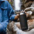 A drink bottle in matte black, featuring a stainless steel rim and a logo, placed on a log near a waterfall.