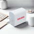 A white travel adapter with a plug and a branding logo, set on a minimalist desk with a keyboard and cup.
