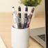 A pen holder featuring several metal pens in white, black, blue, and maroon, placed near a laptop and plant.