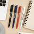Four ballpoint pens in black, blue, red, and white arranged on a desk beside a notebook and laptop.