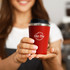A red coffee cup with a silver lid, held by a person, features a logo on the front.