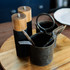 Keepsake Noir salt and pepper grinders in black and wood, alongside utensils in a metal holder on a wooden tray.