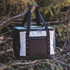 A cooler bag in grey and black with sturdy handles and a front pocket, hanging from a tree branch.