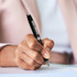 A black ballpoint pen held in a hand, with a person writing on a notepad. The pen has a metallic finish and branding.