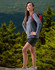 A model wearing a grey long-sleeve tee shirt and black shorts, standing outdoors with a backpack in a wooded area.