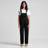 A pair of black canvas overalls worn by a model, paired with a white t-shirt and brown shoes.
